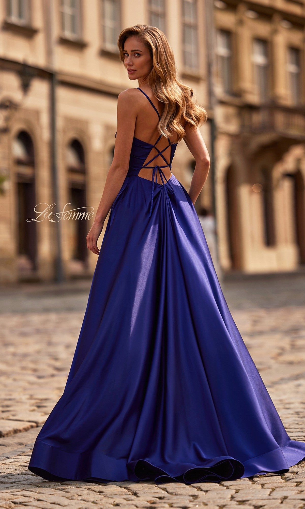 A woman wearing the La Femme 33373 long royal blue satin prom dress with an open strappy back stands on a cobblestone street, glancing over her shoulder.